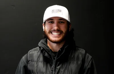 A man with a beard wearing a white baseball cap, black puffer vest, and black hoodie smiles in front of a plain dark background.