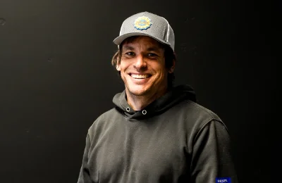 A man in a gray baseball cap and black hoodie smiles while standing against a plain dark background.