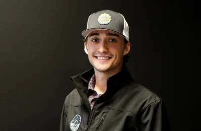 A man wearing a dark jacket and a gray cap with a colorful patch stands against a plain black background, smiling at the camera.