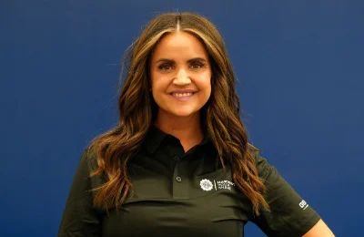 A woman with long brown hair, wearing a black collared shirt with a logo, stands and smiles in front of a plain blue background.