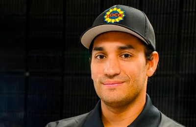 A man wearing a black collared shirt and a black cap with a colorful embroidered logo stands in front of a dark background.