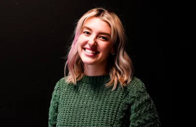 A person with shoulder-length blonde hair wearing a green knit sweater is smiling in front of a plain dark background.
