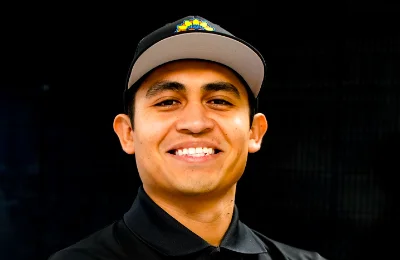 A man wearing a black collared shirt and a cap smiles at the camera against a dark background.