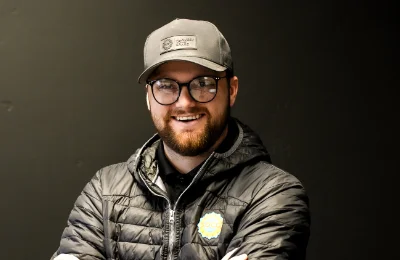 A man wearing glasses, a gray cap, and a black puffer jacket stands against a plain dark background, smiling with his arms crossed.
