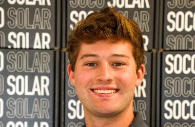 A young man with short brown hair smiles in front of a wall with repeated "SOCIAL SOLAR" text.