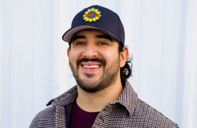 A man with a beard and mustache smiles at the camera, wearing a plaid jacket and a black cap with a colorful circular logo, standing in front of a white corrugated background.