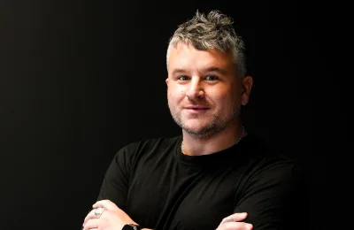 A man with short gray hair and a beard wears a black t-shirt and smartwatch, standing against a black background with his arms crossed.