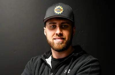A man with a beard wearing a black jacket and a black baseball cap with a colorful circular logo stands against a dark background.