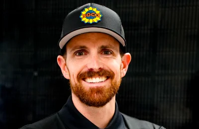 A man with a beard and mustache, wearing a black cap with a colorful logo and a black collared shirt, smiling at the camera against a dark background.