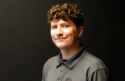 A person with short curly hair wearing a gray collared shirt stands against a plain dark background, looking at the camera and smiling slightly.