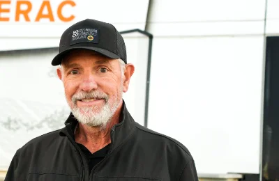 An older man with a gray beard wearing a black cap and jacket stands outdoors, smiling at the camera. The background includes part of a sign with orange letters.