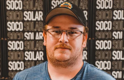 A person wearing glasses, a dark cap, and a blue shirt stands in front of a wall with repeating "SOCO SOLAR" logos.