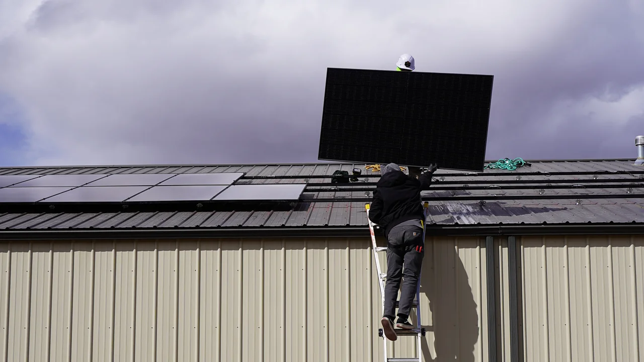 A worker on a ladder installs a solar panel on the roof of a metal building under a cloudy sky.