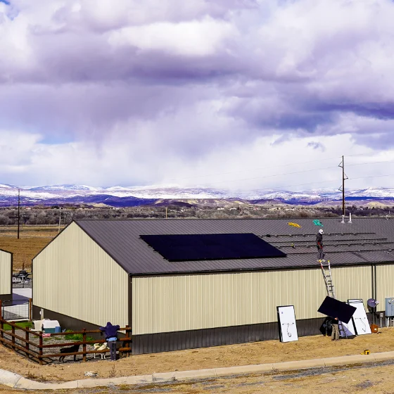 Workers install solar panels on the roof of a large beige industrial building in a rural area with mountains and a cloudy sky in the background.