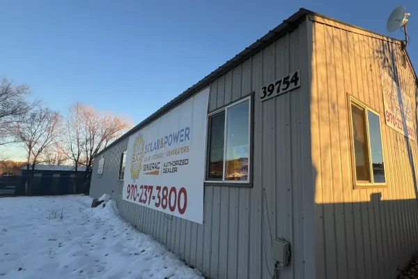 A gray building with windows and a large "Solar Power" banner displaying a phone number, situated in a snowy area with leafless trees and a satellite dish on the roof.