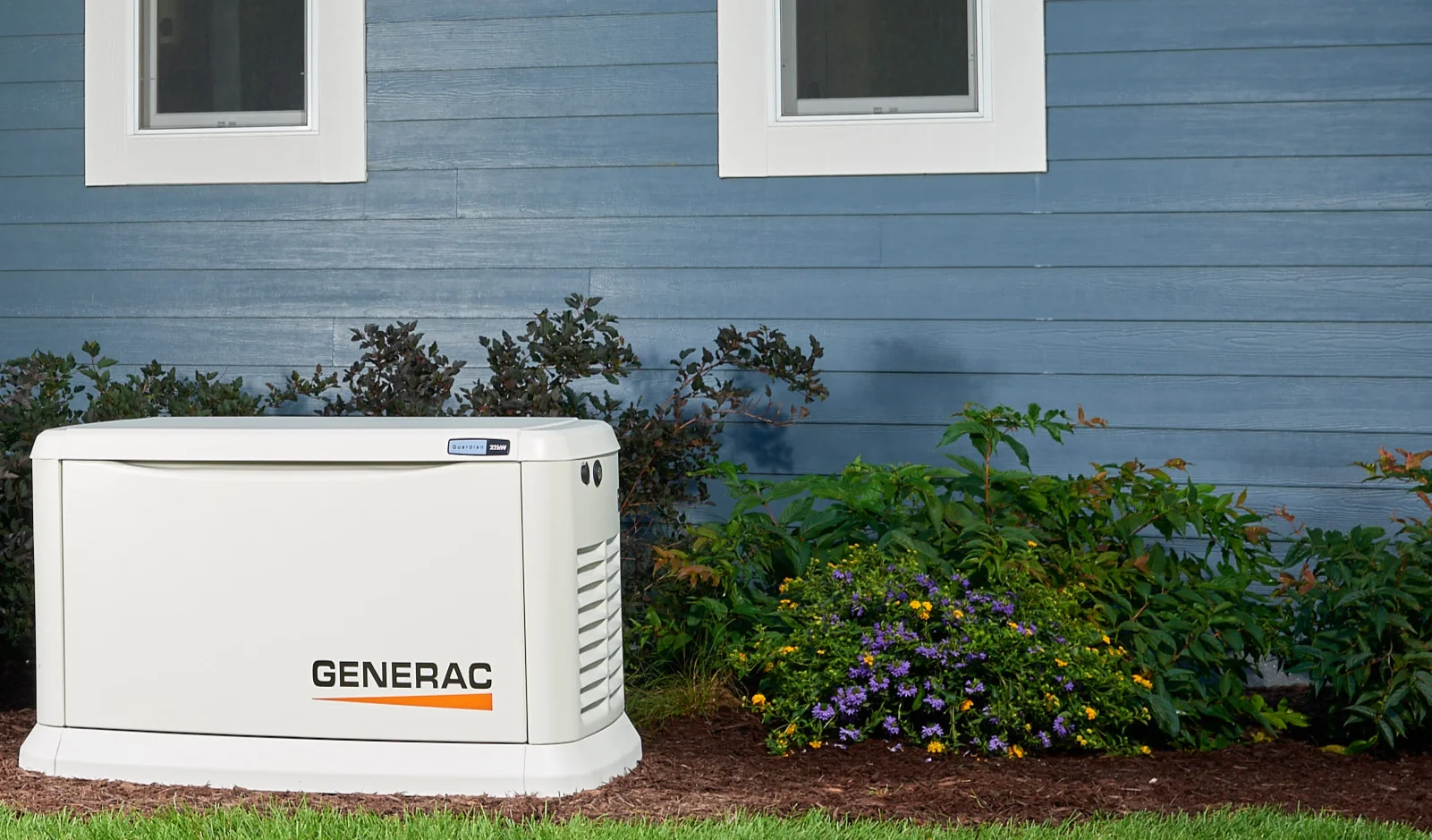 A Generac standby generator is installed outside a blue house with white-trimmed windows and surrounded by shrubs and flowers.