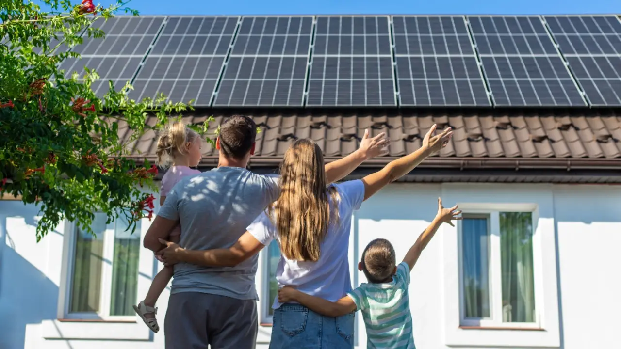A family of four stands in front of a house with solar panels on the roof, pointing and looking up at them on a sunny day.