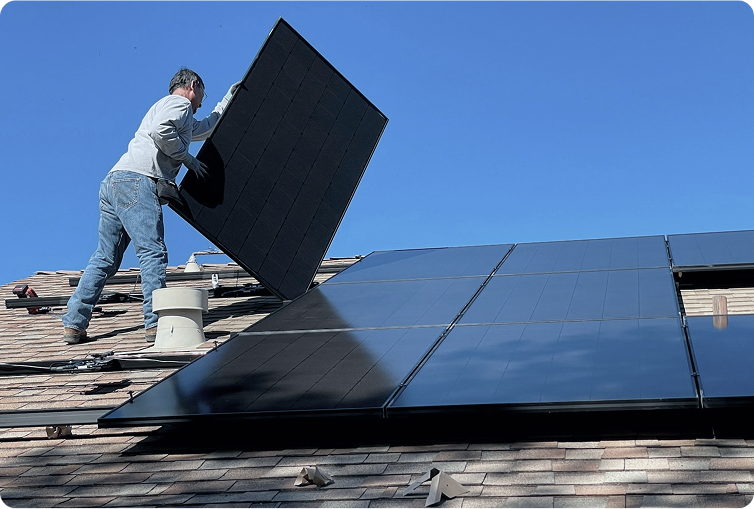 A person installs a solar panel on a residential roof alongside several already-mounted panels under a clear blue sky.