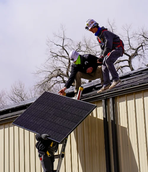 Three workers install a solar panel on the roof of a metal building. Two are on the roof guiding the panel, while one lifts it from below. Bare trees are visible in the background.