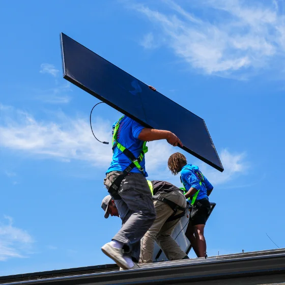 Three workers in safety gear install a solar panel on a rooftop under a blue sky with light clouds.