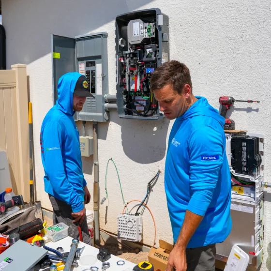 Two technicians in blue hoodies work on electrical panels mounted on an exterior wall, surrounded by tools and equipment.