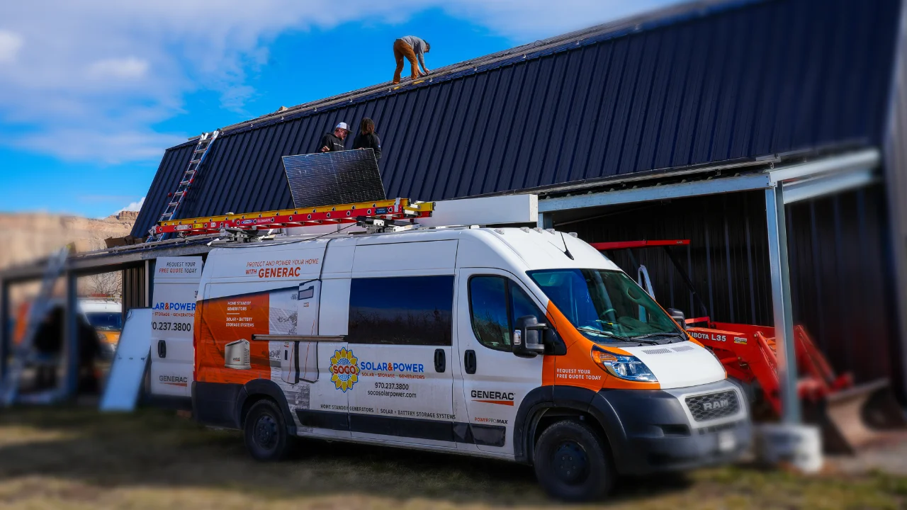 A solar power company van is parked beside a building while two workers install solar panels on the roof using ladders and equipment.