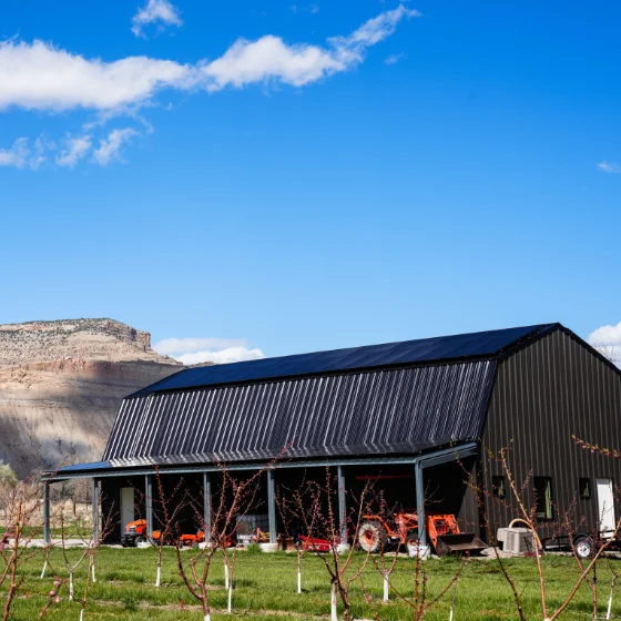 A black metal barn with a sloped roof sits on green grass, with farming equipment outside and rocky hills in the background under a clear blue sky.