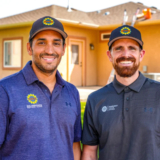 Two men wearing branded polo shirts and caps stand outside in front of a house, smiling at the camera.
