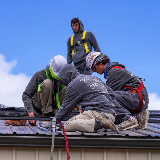 Three workers in safety gear install equipment on a metal roof while another person stands behind them, all under a blue sky.