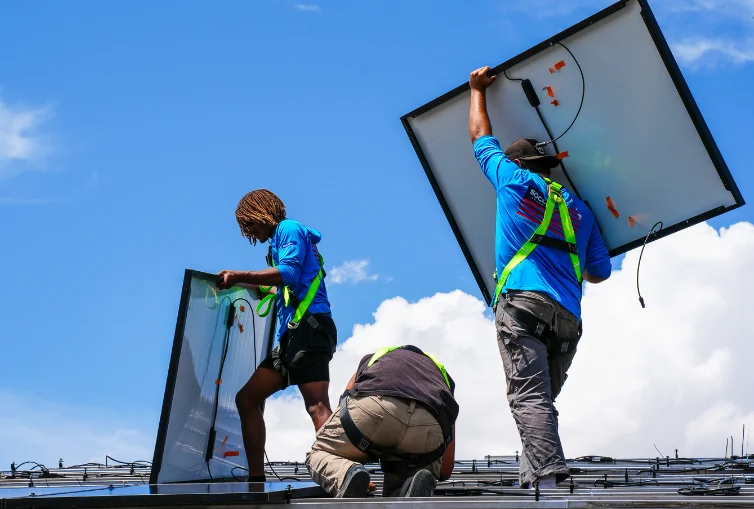 Three workers wearing safety harnesses install solar panels on a rooftop under a bright blue sky with clouds.