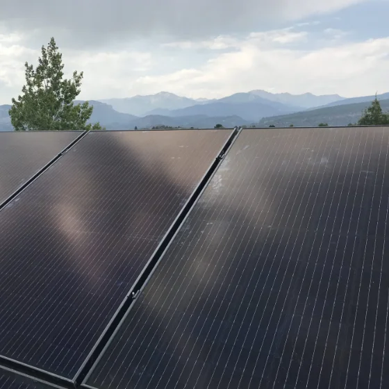 Solar panels are installed outdoors with mountains and trees visible in the background under a cloudy sky.