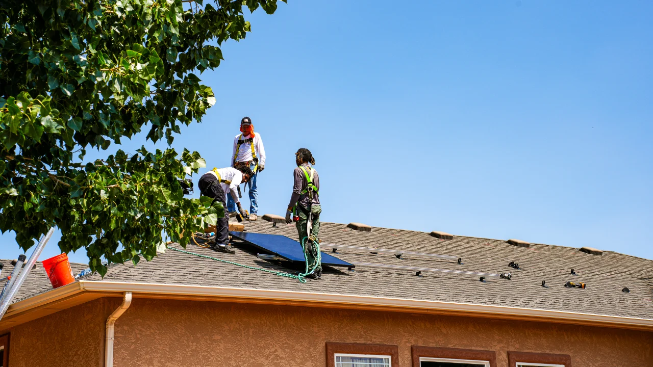 Three workers install solar panels on the roof of a house under a clear blue sky, with a tree partially visible on the left side.